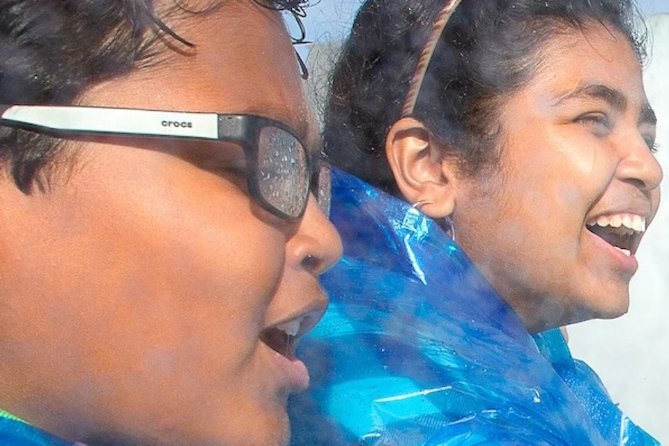 Children wearing blue rain ponchos smile while standing on a boat near a large waterfall and a visible rainbow.