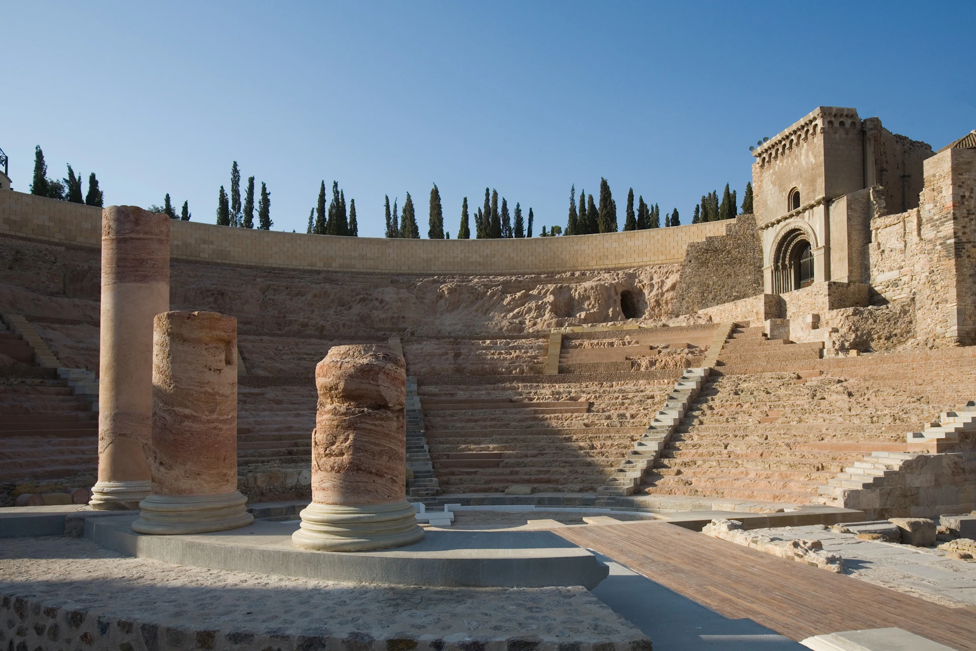 Ancient amphitheater with stone steps, columns in the foreground, and cypress trees on the horizon. Clear blue sky.
