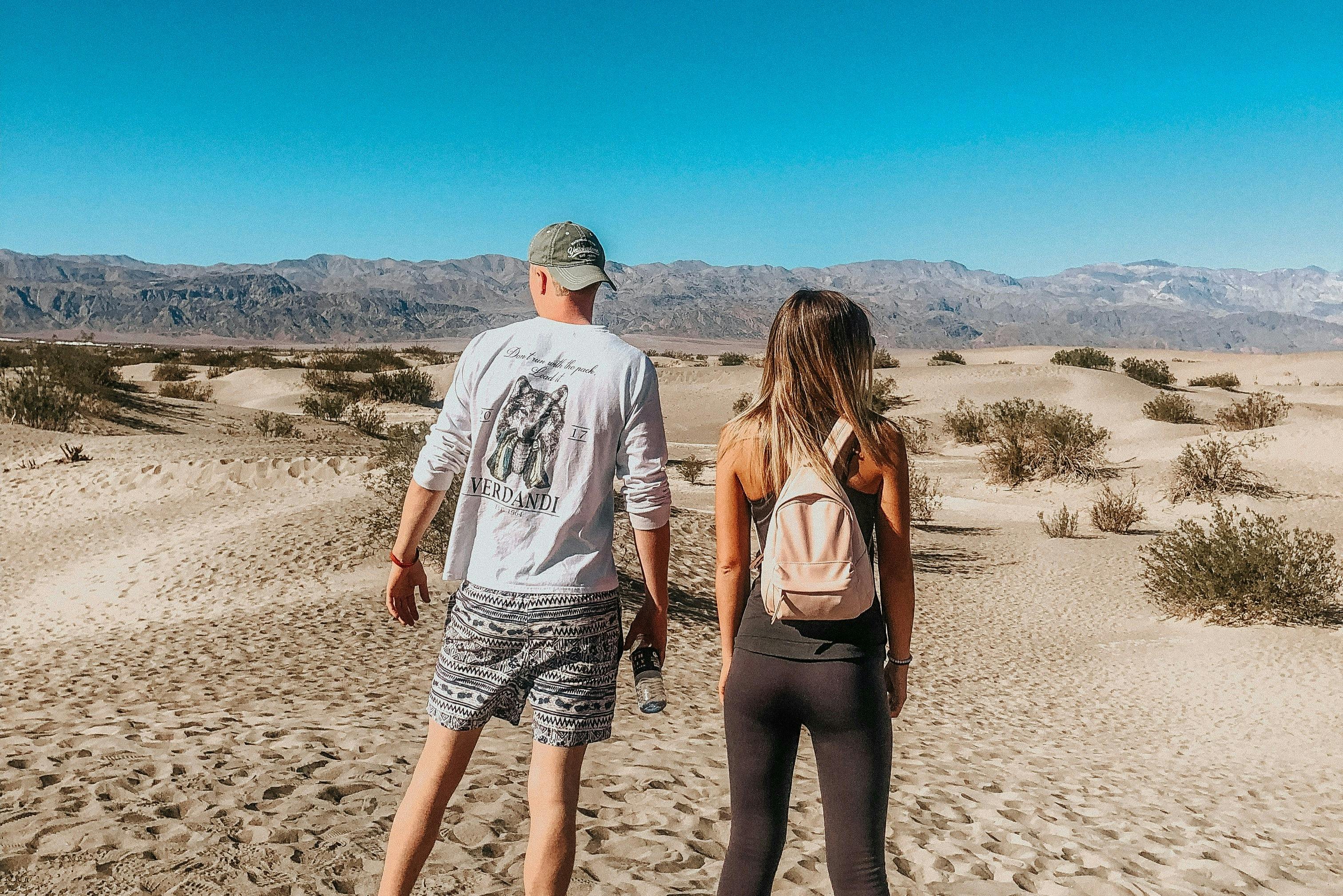 Two people walking in a desert landscape with sandy terrain, sparse vegetation, and mountains in the distance.