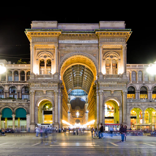 Upplyst neoklassisk byggnad med välvd entré på natten, människor som sitter och går, cyklar närvarande.