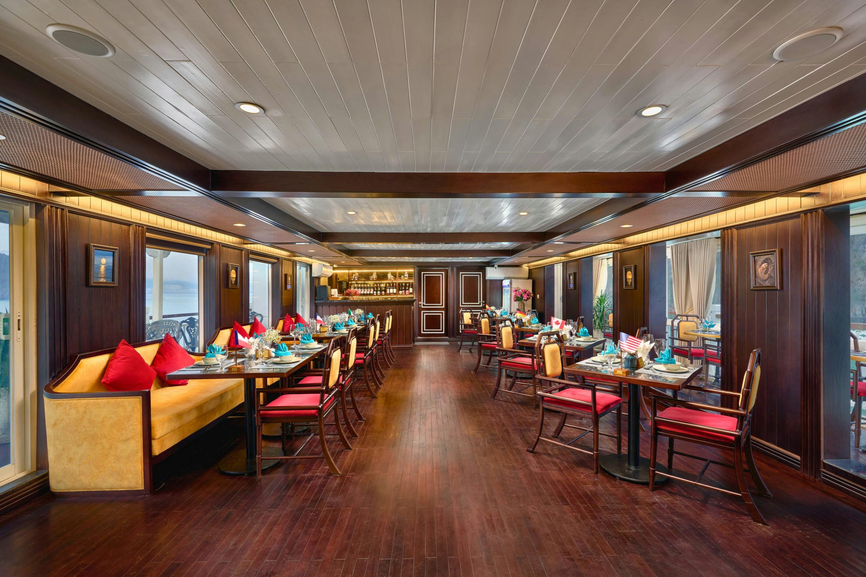 A spacious restaurant with wooden floors, set tables, red cushioned seating, and large windows along the sides.