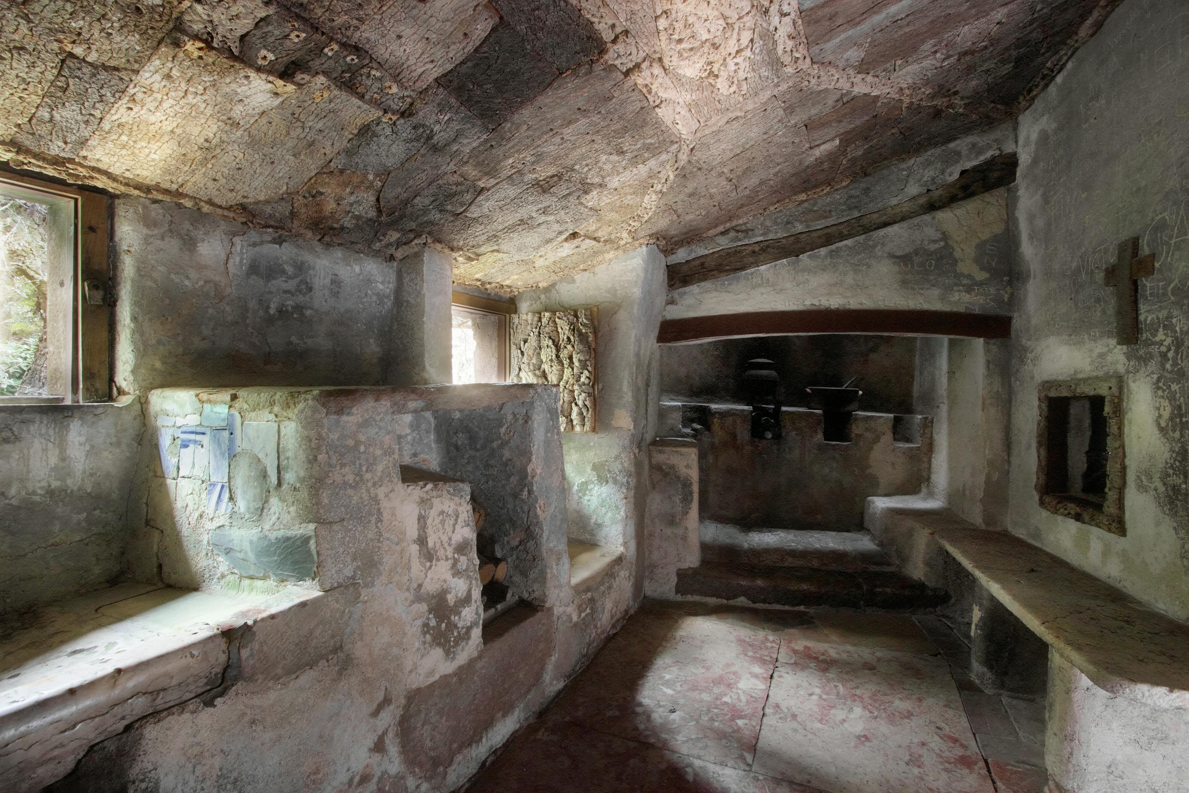 Una vecchia stanza in pietra con un soffitto inclinato, pareti grezze e una piccola finestra. La stanza contiene scaffali e nicchie in pietra.