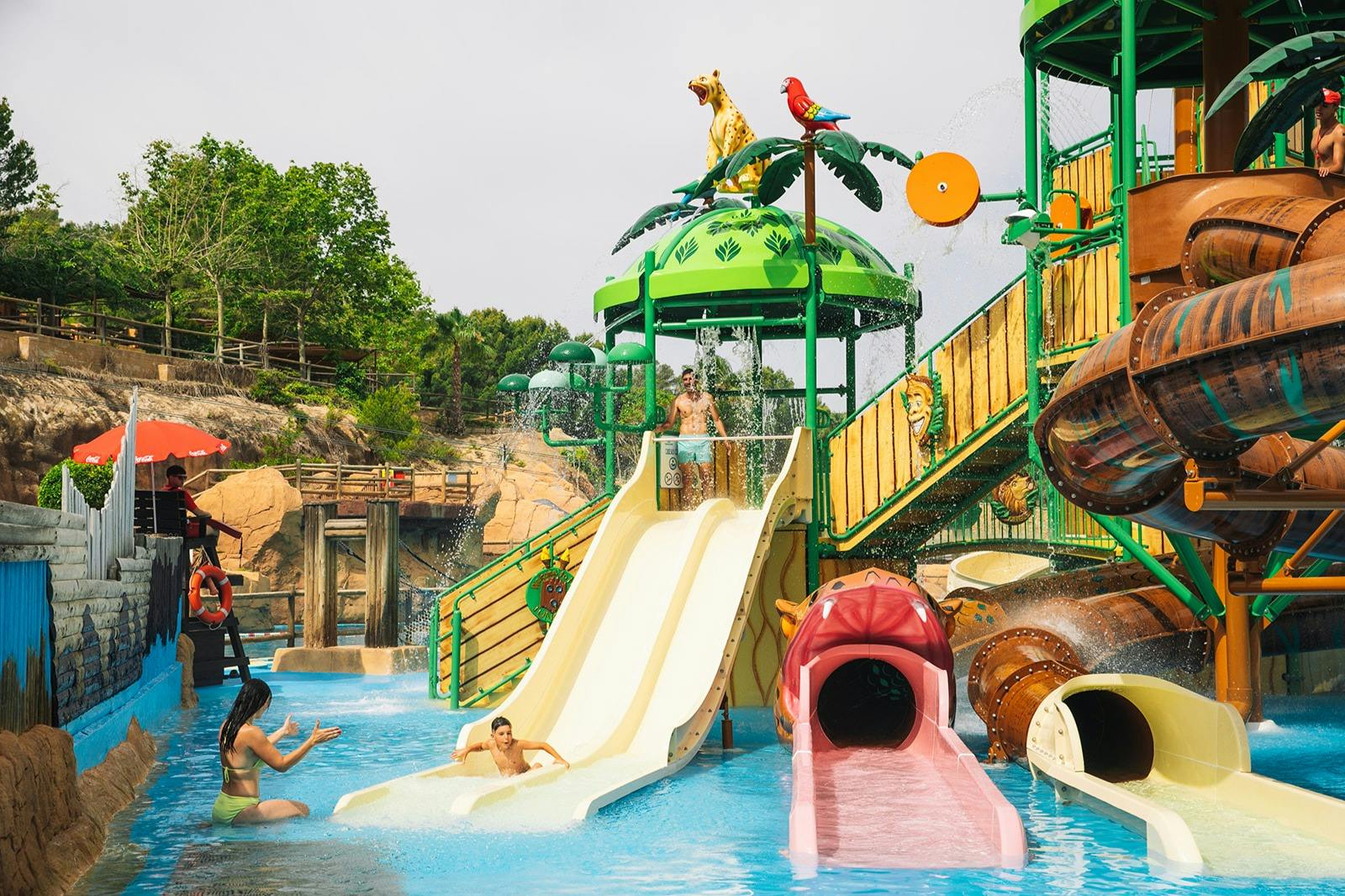 Un divertente parco acquatico con scivoli colorati, un’area giochi d’acqua e gente che si diverte in acqua sotto un cielo soleggiato.
