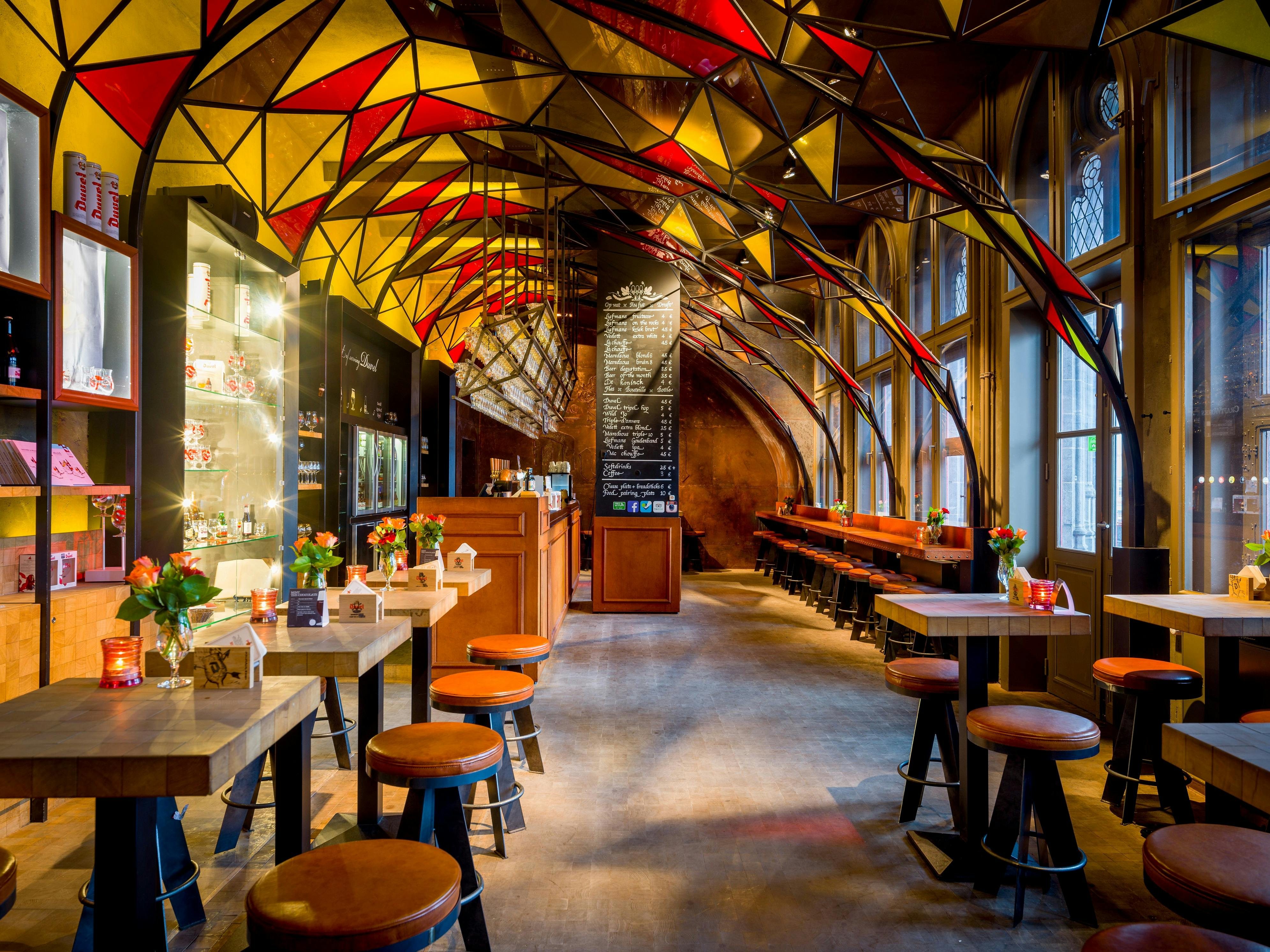 Stylová kavárna s barevným geometrickým stropem, dřevěnými stoly a stoličkami, barovým pultem, jídelním lístkem a květinami na stolech.