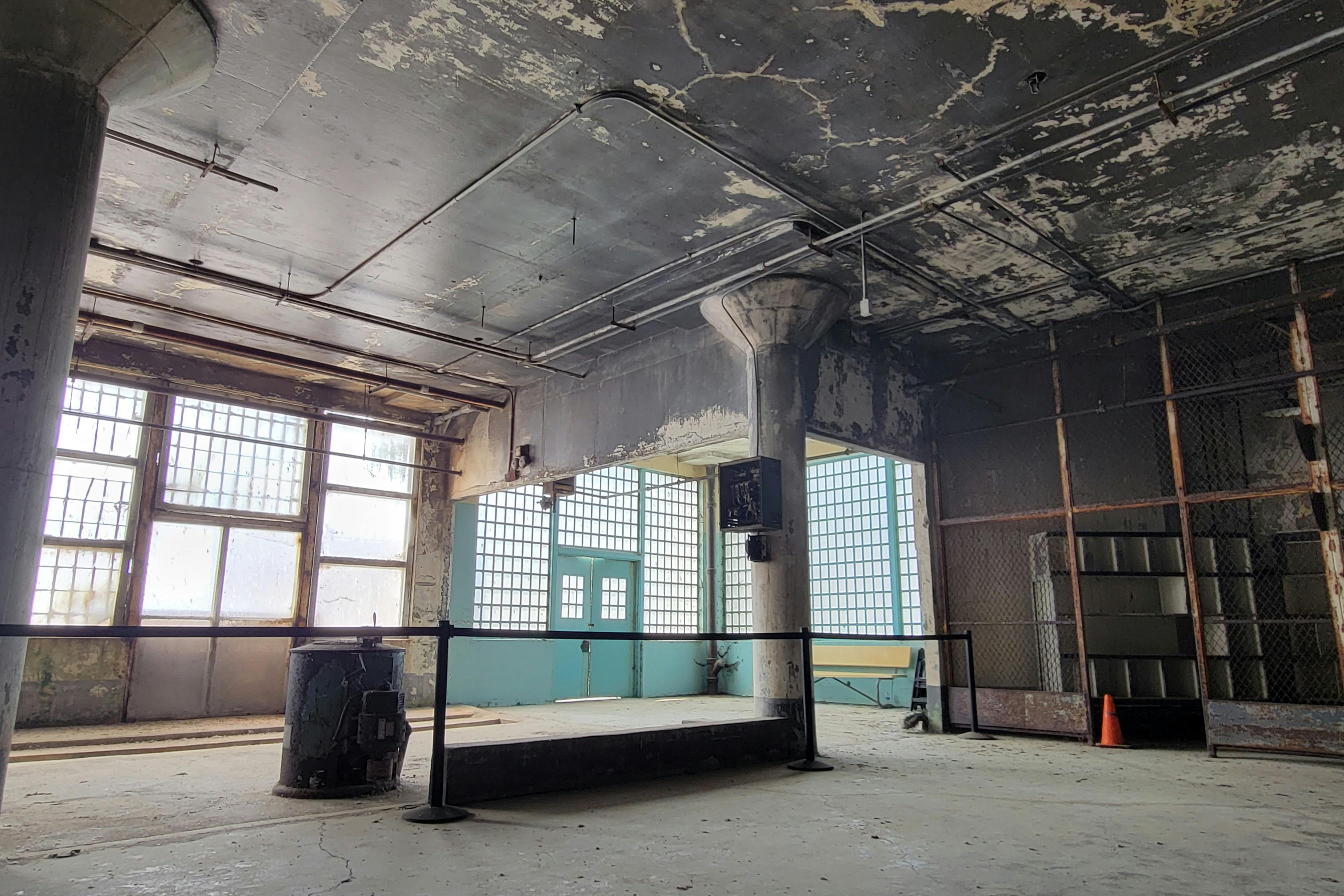 A large, industrial-style room with peeling paint, large windows, a column, and a metal railing across the floor.
