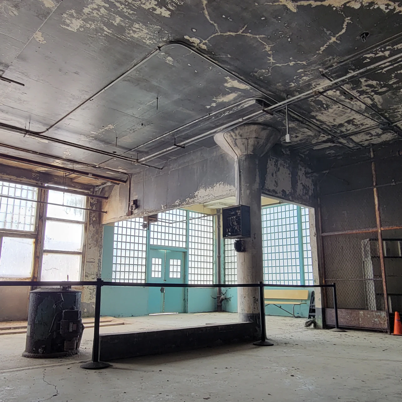 A large, industrial-style room with peeling paint, large windows, a column, and a metal railing across the floor.