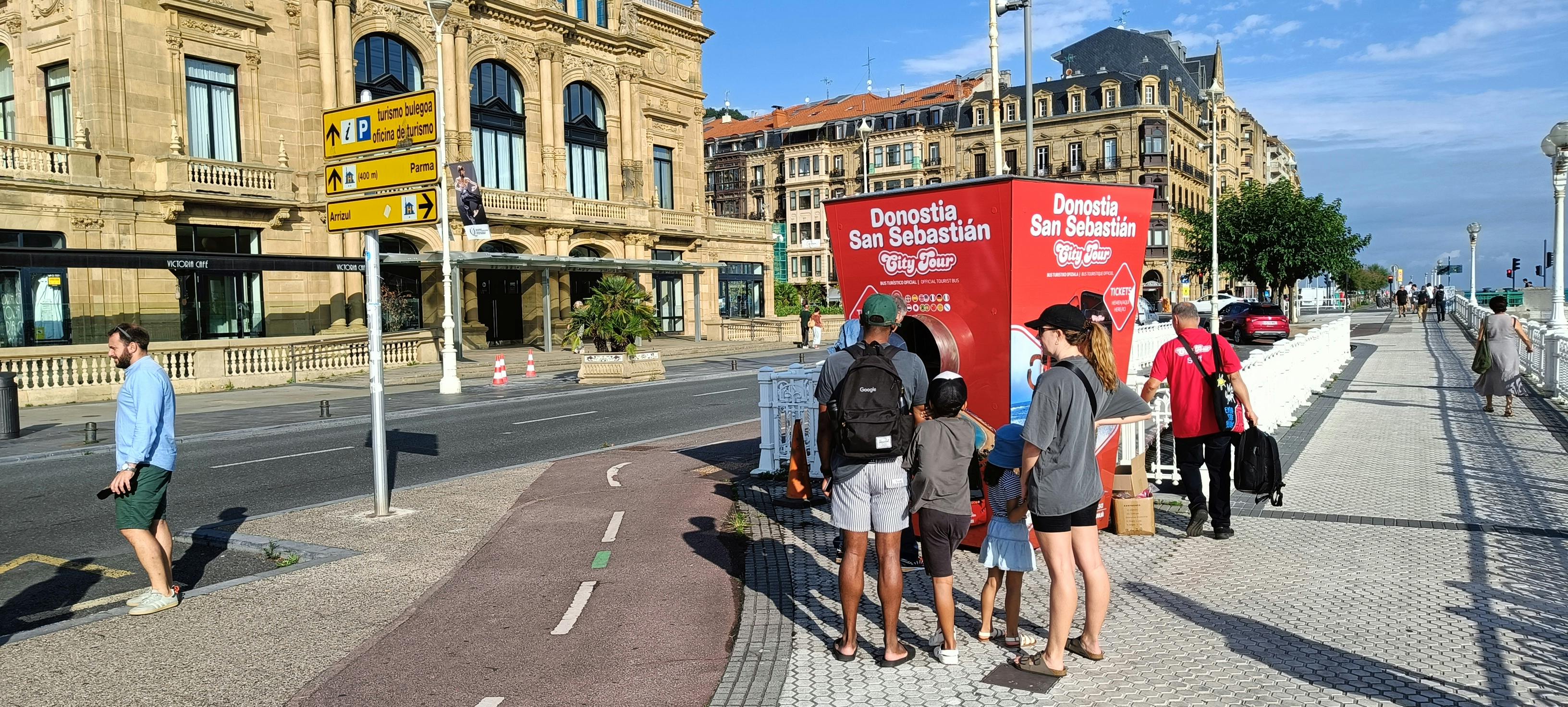 A family examines a red Donostia San Sebastian City Tour kiosk on a sunny street near historic buildings.