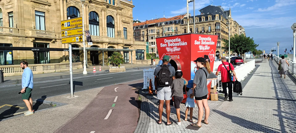 A family examines a red Donostia San Sebastian City Tour kiosk on a sunny street near historic buildings.