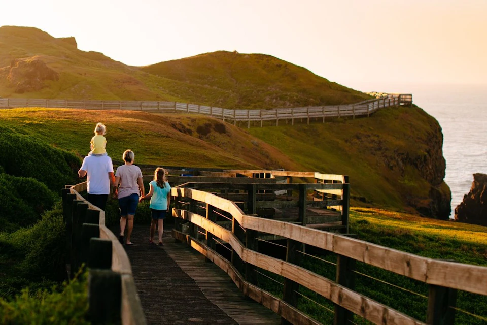 A family walking on a wooden boardwalk through grassy hills at sunset, with a child on someone's shoulders.