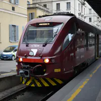 Un treno marrone con la scritta "Malpensa T2" in una stazione urbana con edifici e auto parcheggiate sullo sfondo.