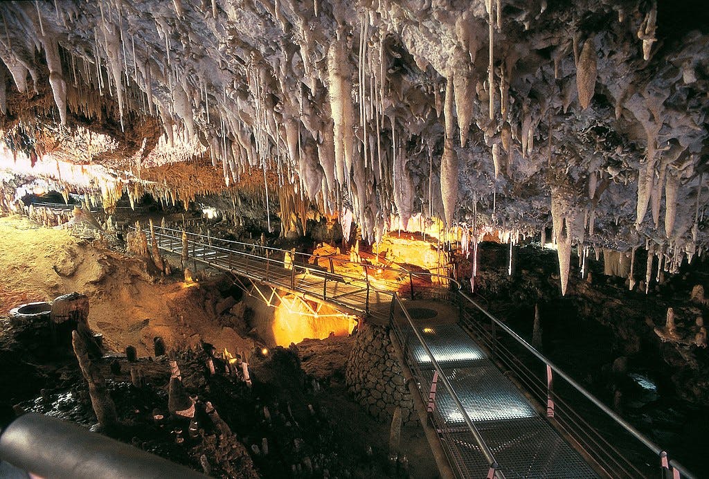 Grotta med stalaktiter och stalagmiter, upplyst gångväg med räcken och ett glödande orange område som belyser formationerna.