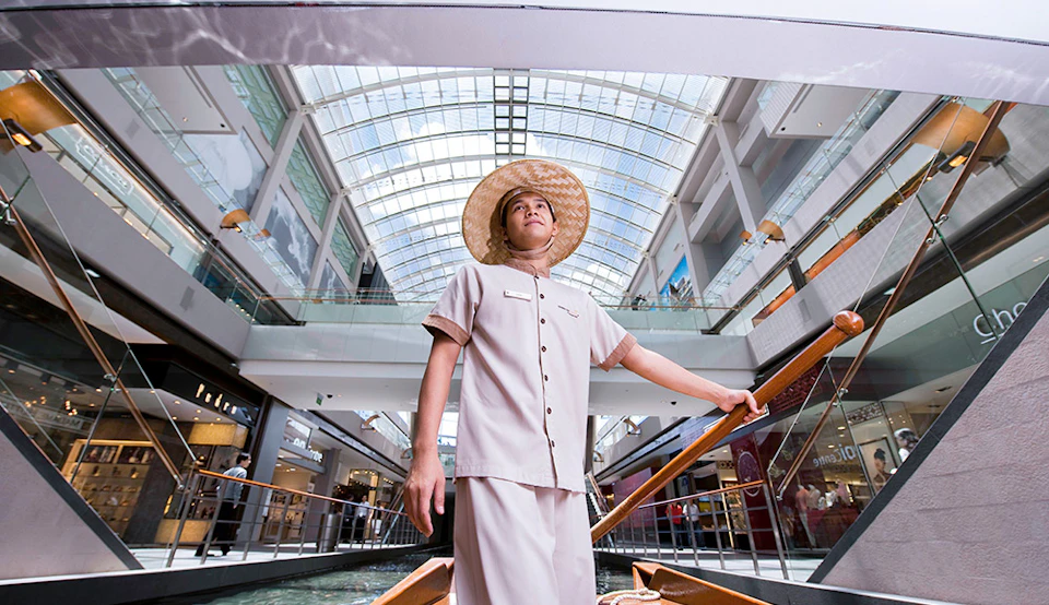 A man in a beige uniform and straw hat stands in a boat inside a modern, glass-roofed shopping mall.