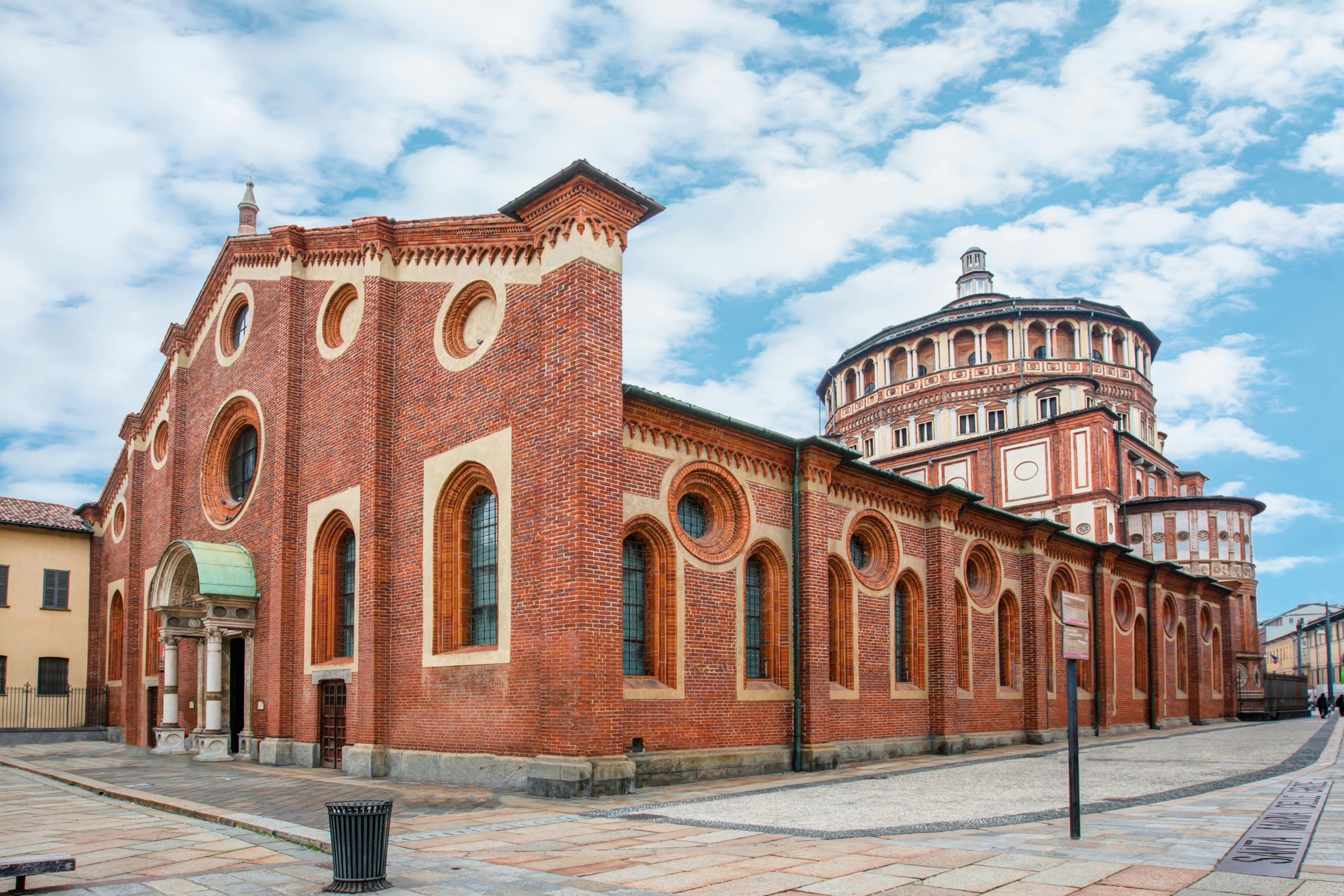 Здание из красного кирпича с арочными окнами и круглыми конструкциями, примыкающее к округлой, богато украшенной конструкции. Голубое небо с облаками над головой.