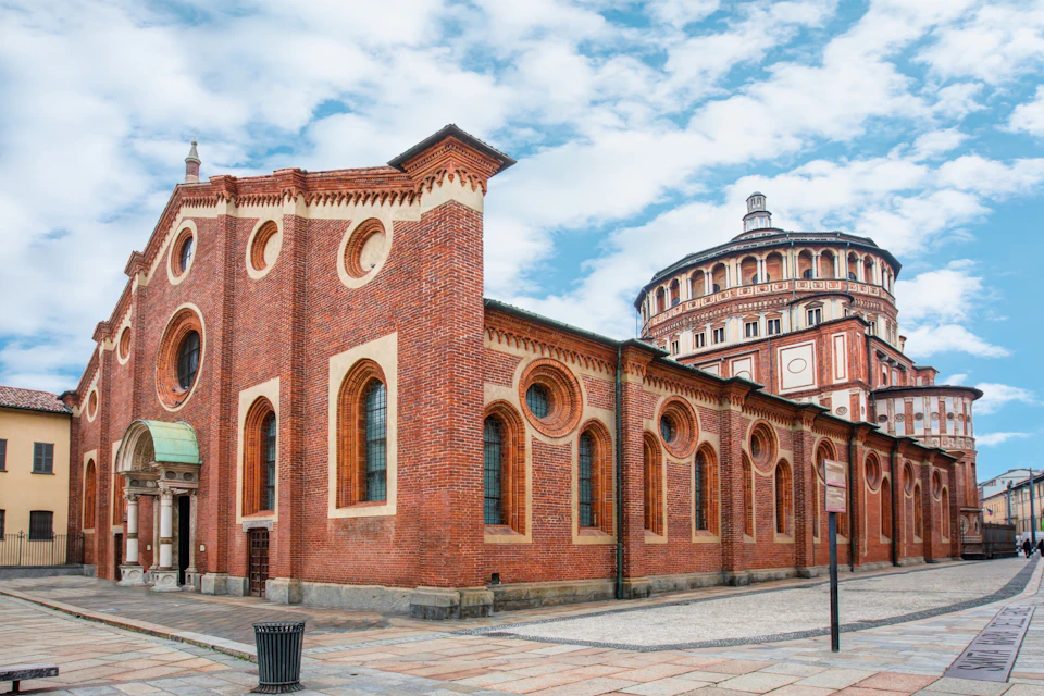 Red-brick building with arched windows and circular designs, adjacent to a rounded, ornate structure. Blue sky with clouds overhead.