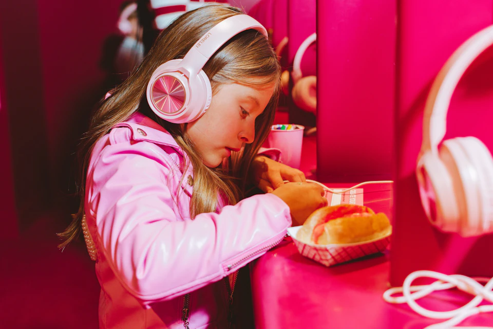 A girl wearing pink headphones and a pink jacket writes at a table with a hot dog in a paper tray.