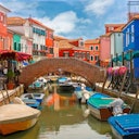 Colorful buildings line a canal with small boats and a brick bridge under a blue sky with clouds.