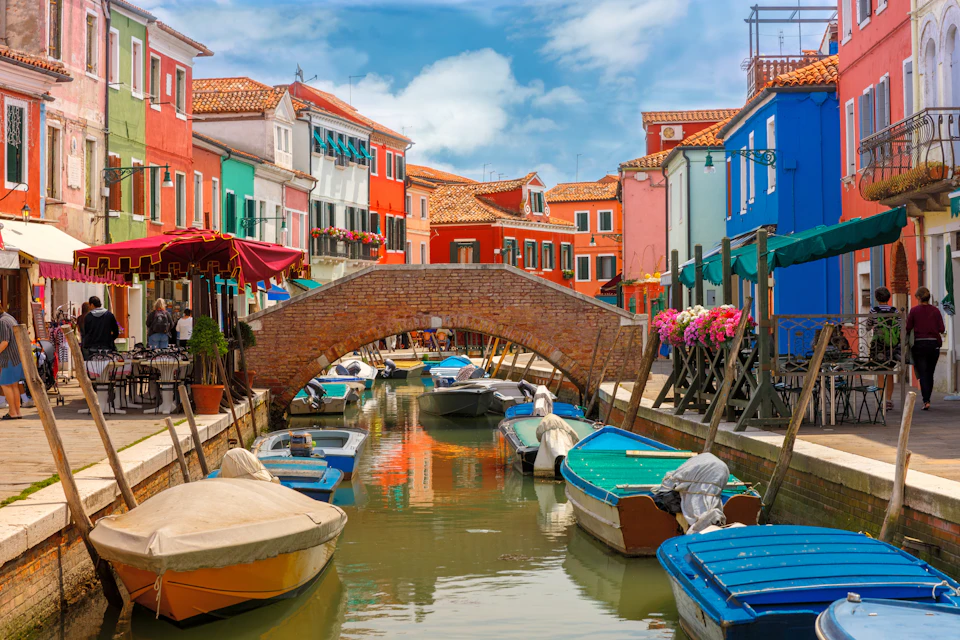 Colorful buildings line a canal with small boats and a brick bridge under a blue sky with clouds.