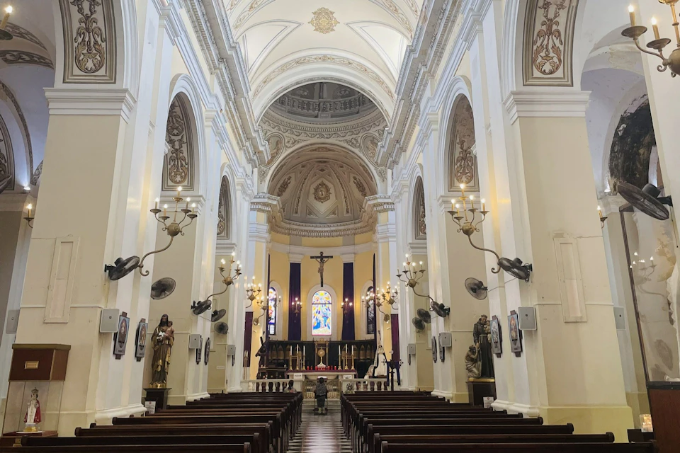 Interior of an ornate church with high arched ceilings, chandeliers, wooden pews, and a central altar with a crucifix.