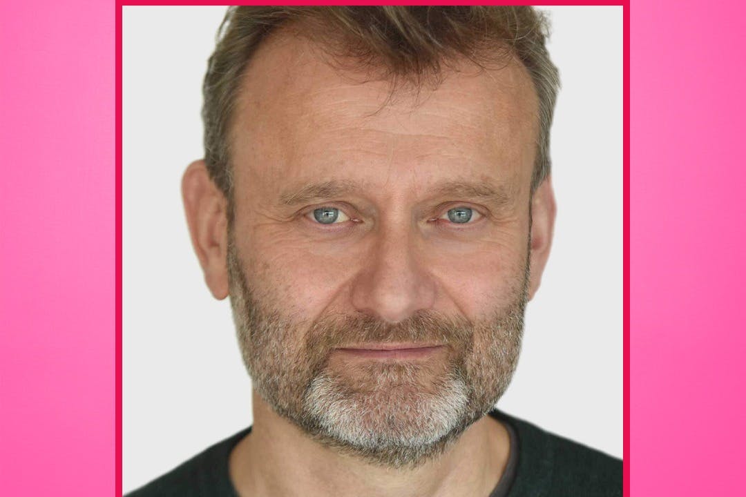 Ένας άντρας με κοντά καστανά μαλλιά και γκρίζα γενειάδα, φορώντας ένα σκούρο πουκάμισο, κοιτάζει απευθείας την κάμερα σε ένα απλό φόντο.