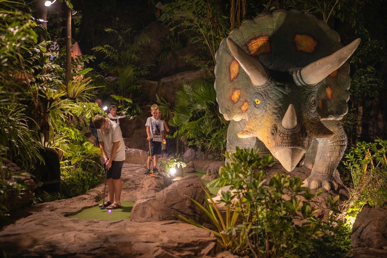 Children playing mini-golf in a jungle-themed setting with a large dinosaur statue and lush plants surrounding the course.