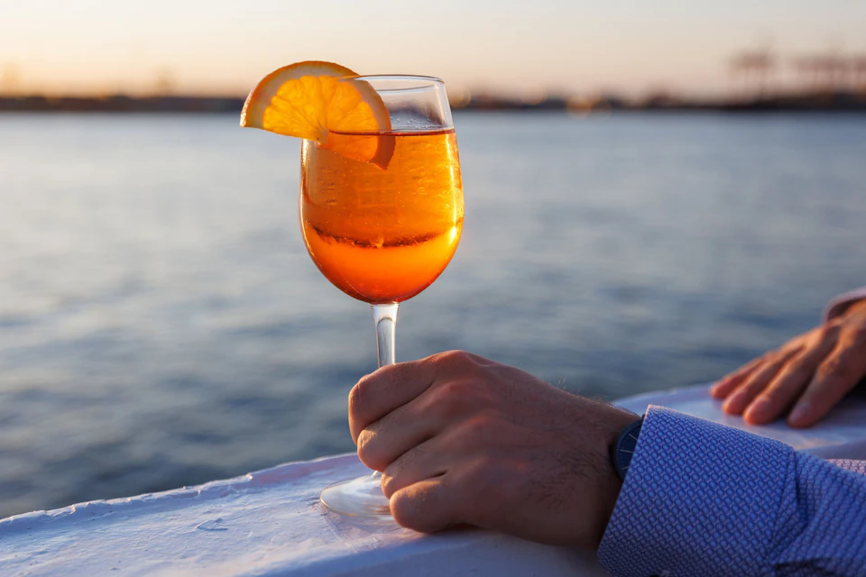 A hand holding a glass of orange cocktail with an orange slice garnish, against a waterfront backdrop at sunset.