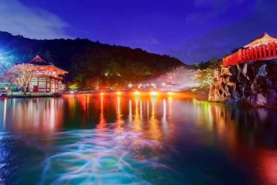 A serene pond reflecting colorful lights and surrounded by trees and a building under a twilight sky.