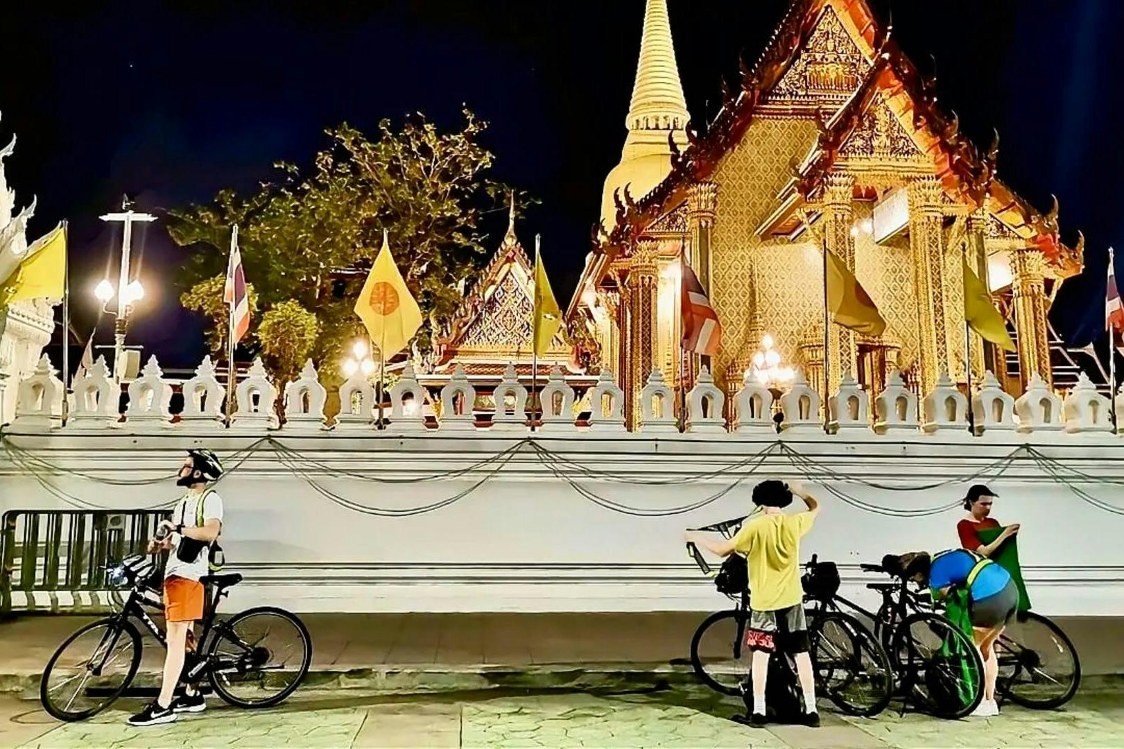 旗や木々に囲まれた明るい華やかな寺院の前に、二人の人が自転車のそばに立っています。