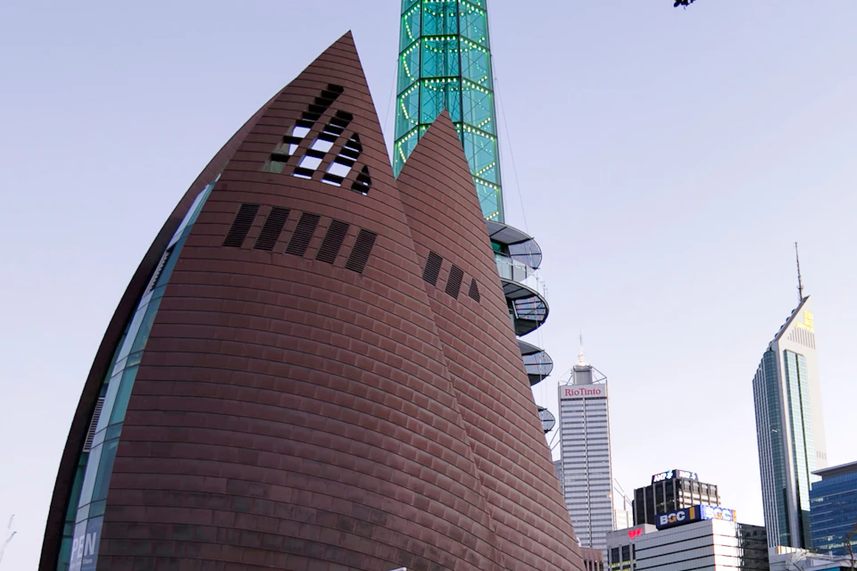 A modern building with triangular shapes, a tall green tower, and city skyscrapers in the background.
