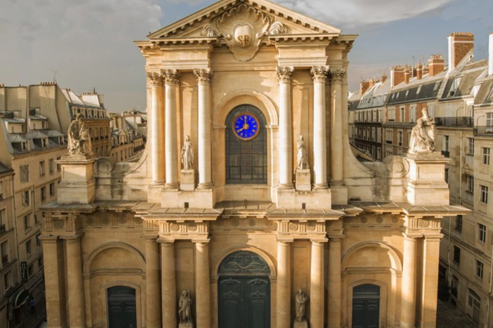 Facade of an ornate historic building with columns, statues, and a blue circular window, framed by a cityscape under a cloudy sky.