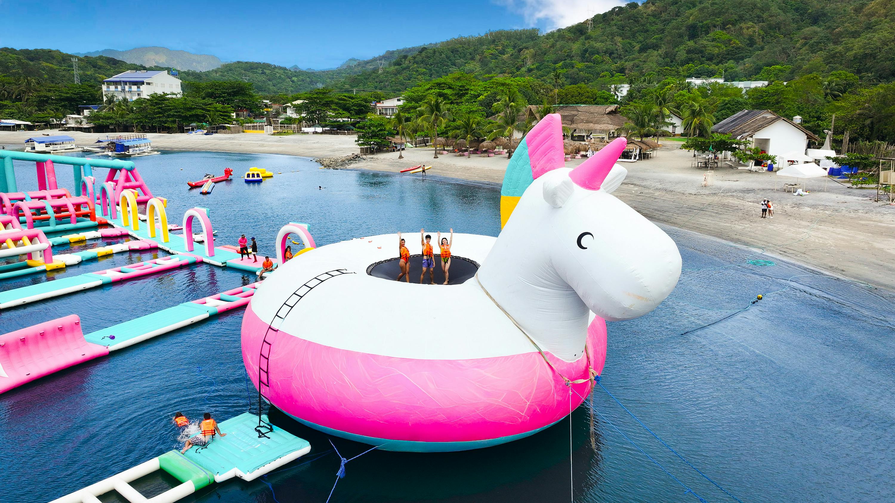 People standing on a large inflatable unicorn float in a water park by the shore, with greenery and buildings in the background.