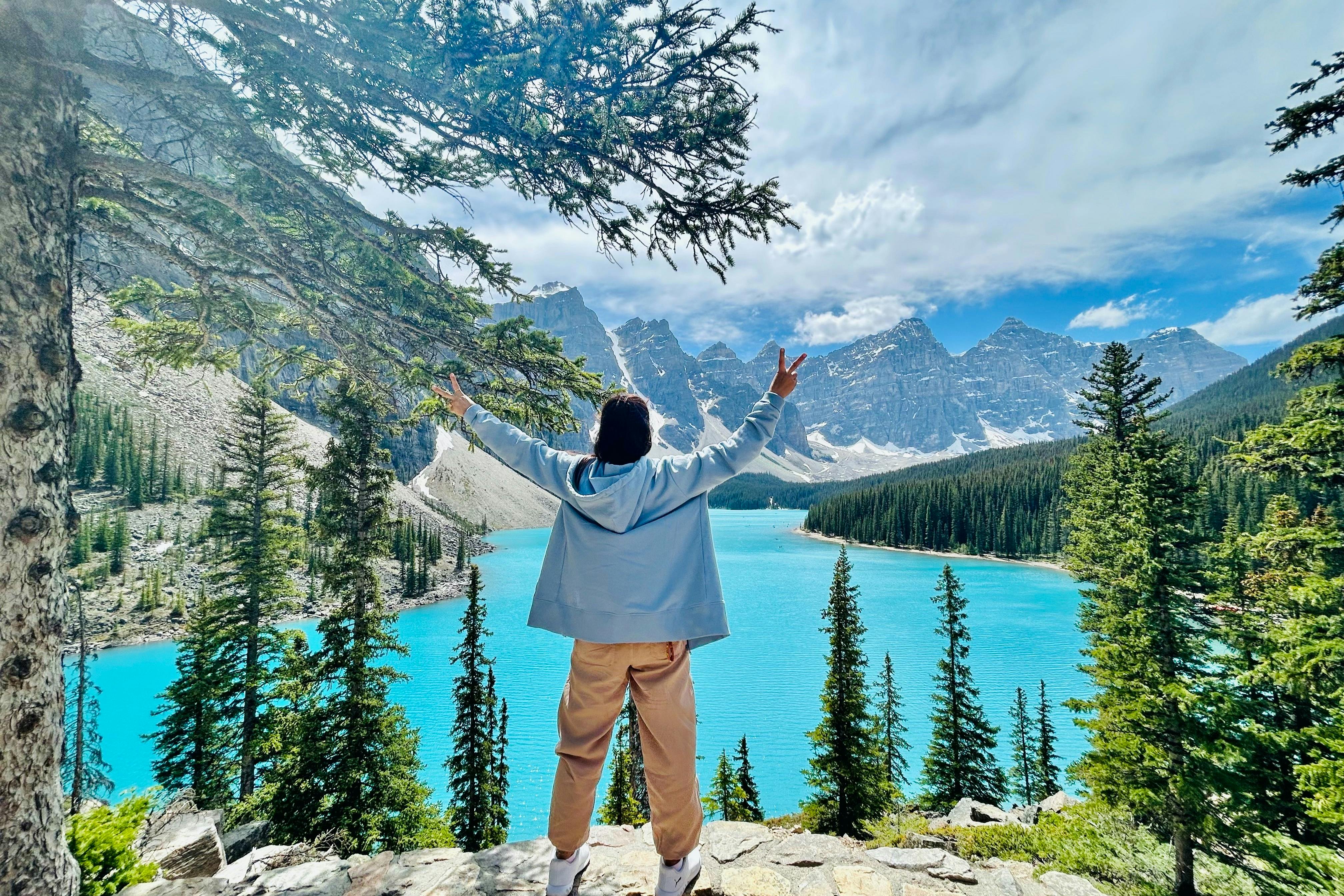 Person with raised arms stands before a turquoise lake, surrounded by pine trees and mountains under a partly cloudy sky.