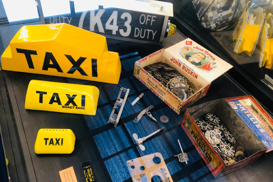 A display of vintage taxi signs, mechanical parts, and boxes, including a yellow taxi money bank and a John Ruskin box.