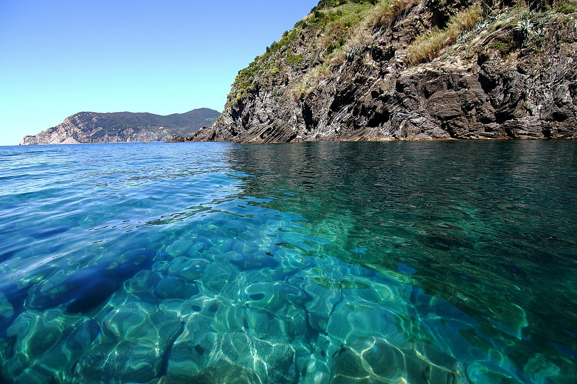 Clear turquoise water with visible rocks below, a rocky coastline with greenery, and distant hills under a clear blue sky.