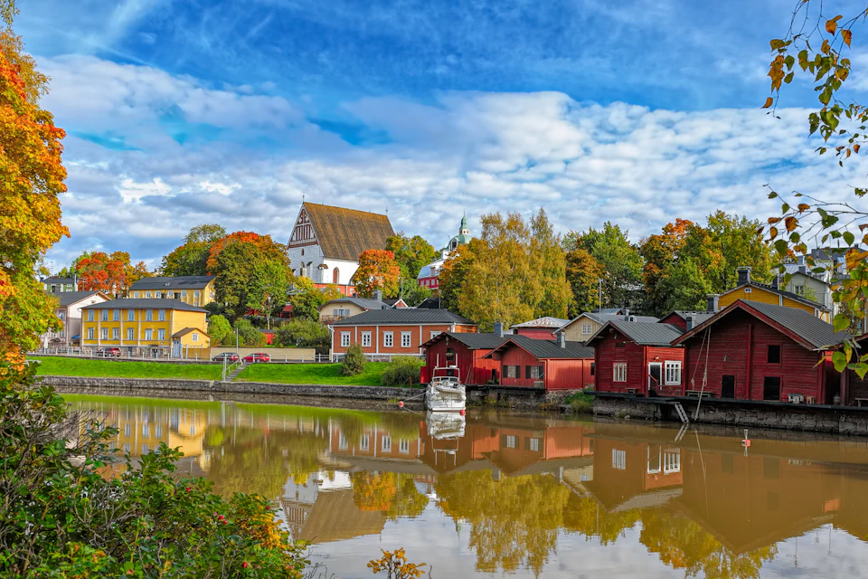 A scenic riverside view with colorful houses, a white church on a hill, autumn trees, and a boat docked along the water.