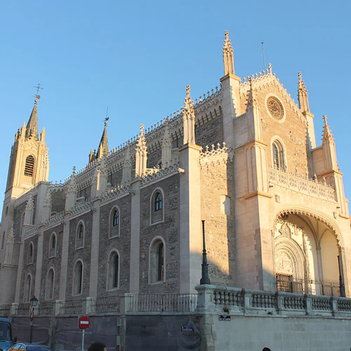 Kyrka i gotisk stil med invecklade stenarbeten, höga spiror, välvd entré och människor som går i närheten under en klarblå himmel.