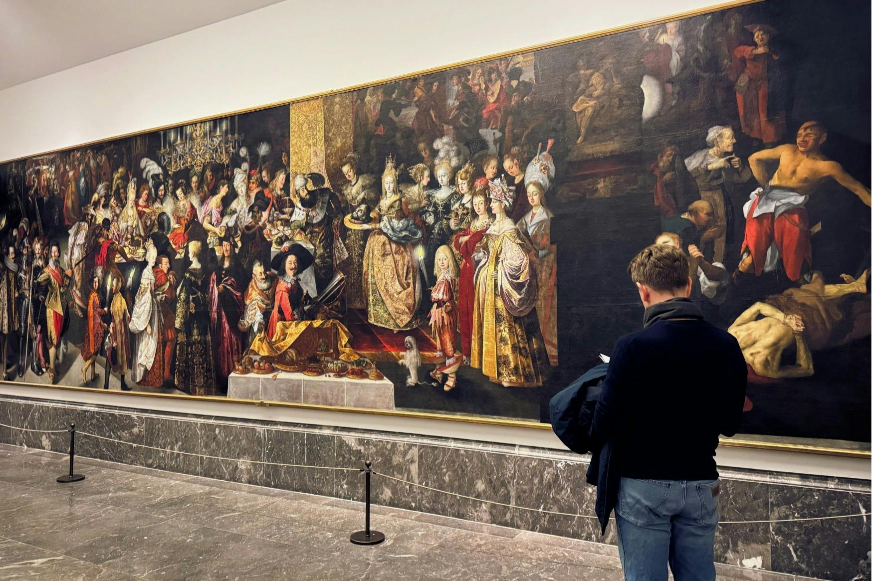 壮大で詳細な歴史的絵画である記念碑的な宮廷宴会の場面を男性が観察しています。