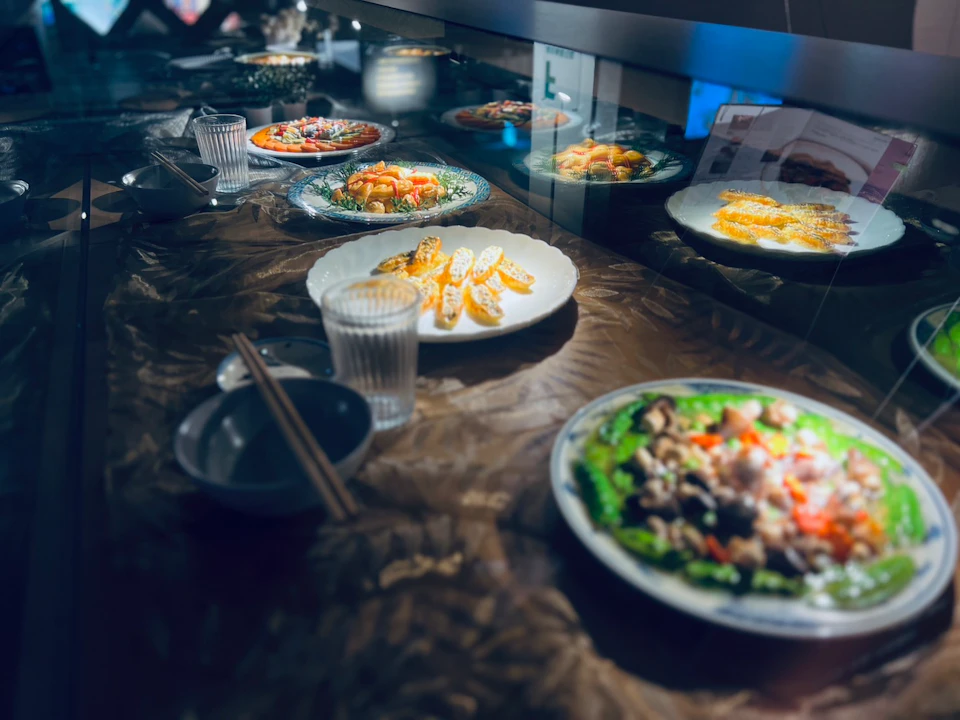 A display of various plated dishes, including vegetables and meats, on a reflective surface with utensils and glasses nearby.