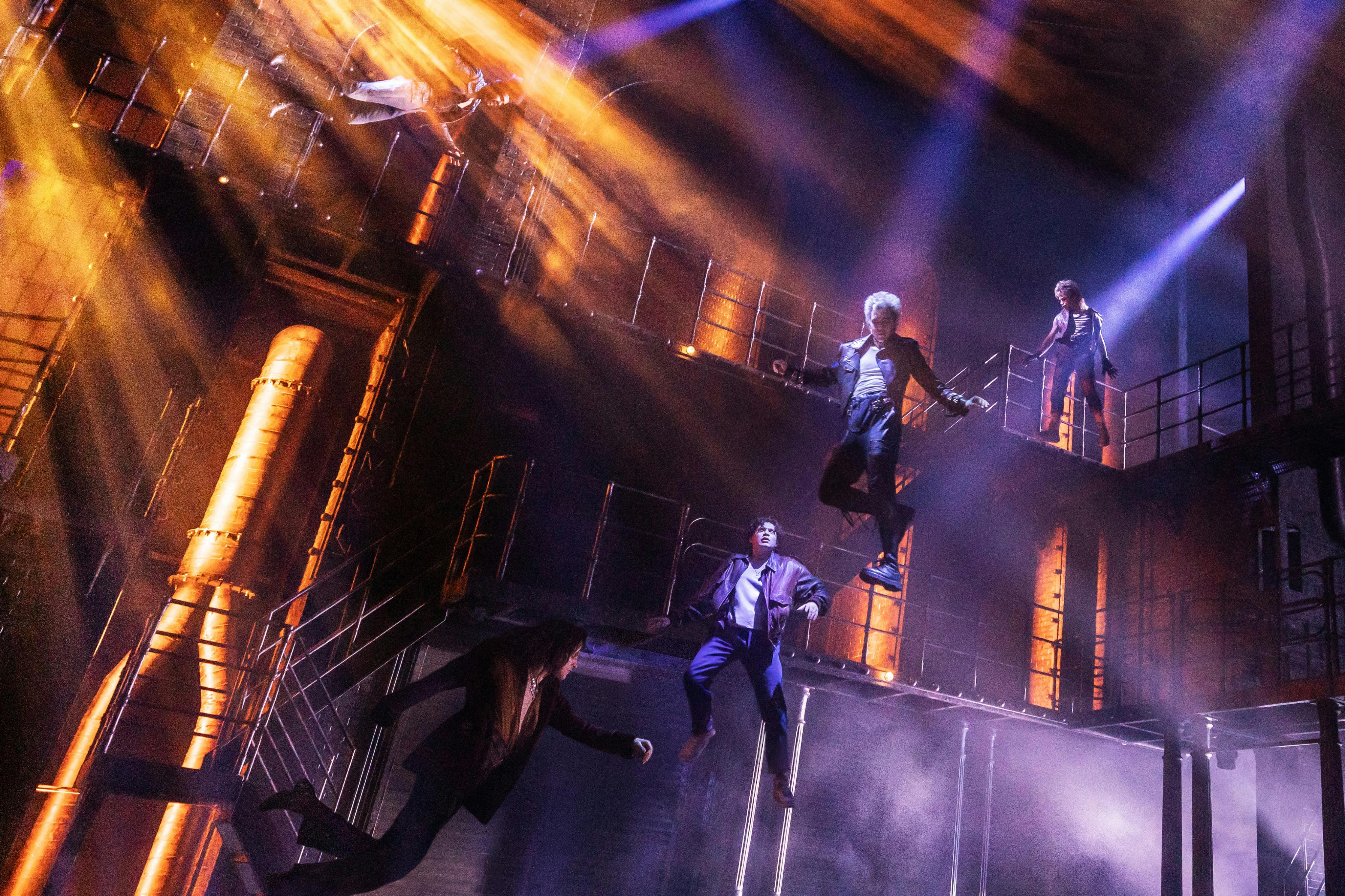 Performers in suits suspended in mid-air on a theatrical stage with dramatic lighting and industrial set design.