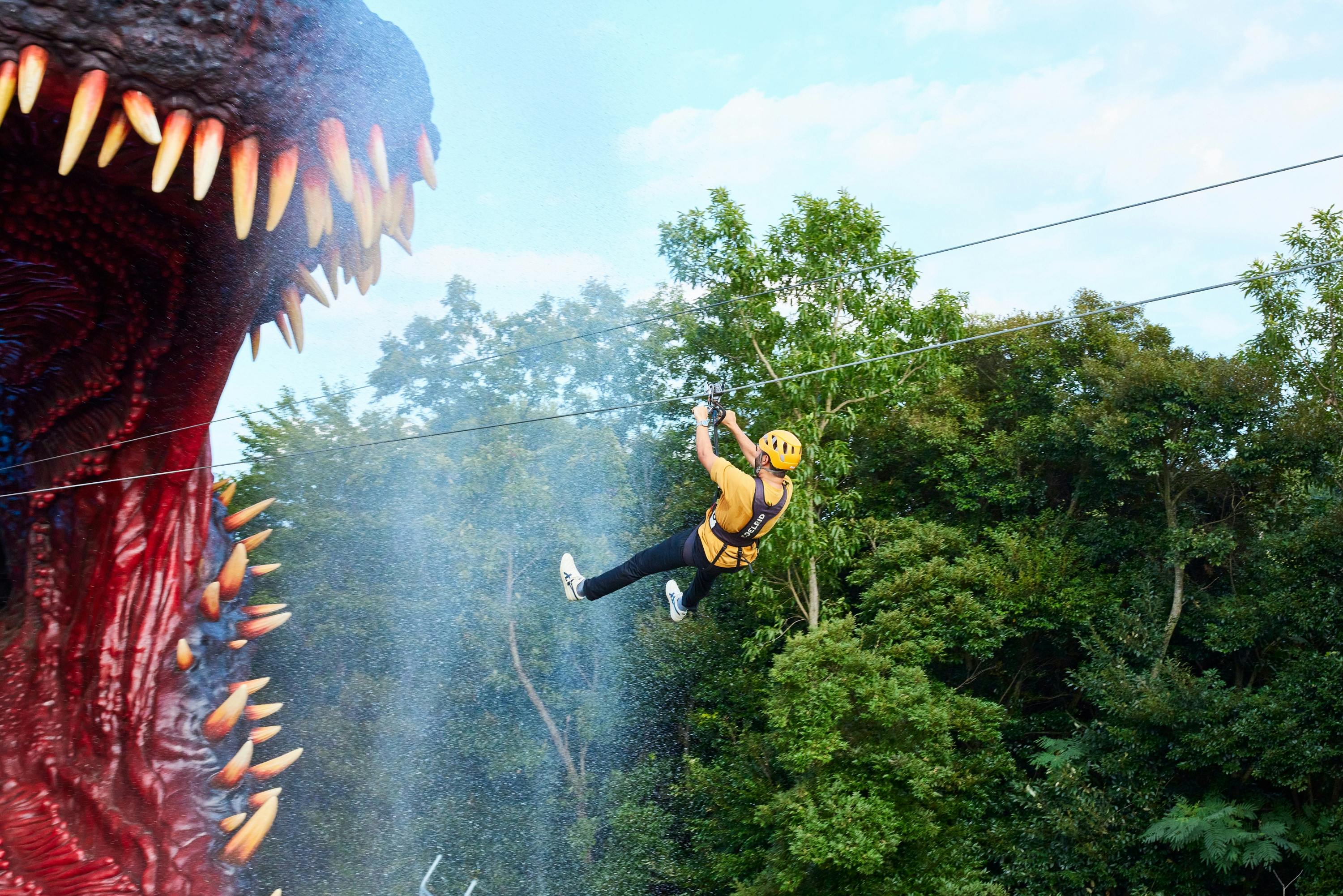 A person ziplining through a forested area, near a large artificial dinosaur head with sharp teeth.