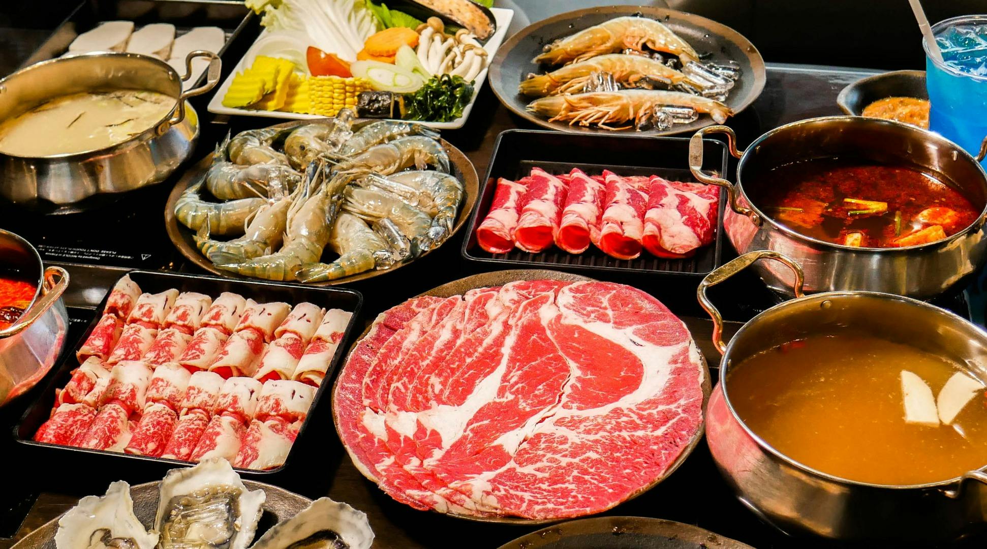 A spread of raw ingredients for hot pot, including sliced meat, shrimp, vegetables, and a pot of broth.