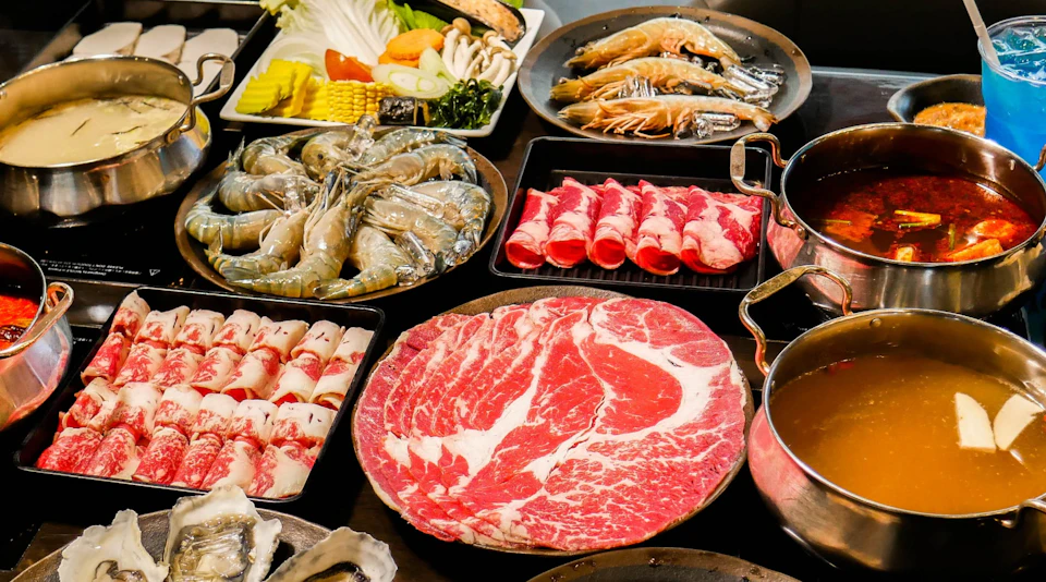 A spread of raw ingredients for hot pot, including sliced meat, shrimp, vegetables, and a pot of broth.