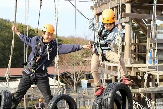 Two people wearing helmets and harnesses participate in an outdoor rope course involving hanging tires and wooden platforms.