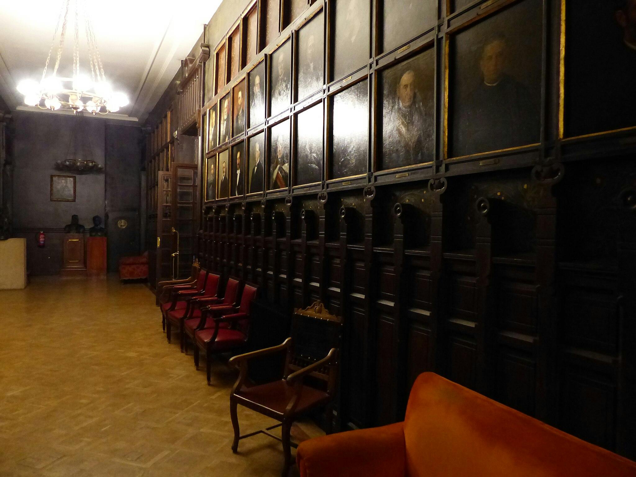 Dimly lit hallway with portraits on the wall, red chairs along one side, chandeliers above, and busts on a far wall.