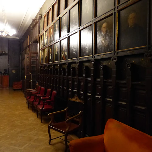 Svagt upplyst hall med porträtt på väggen, röda stolar längs ena sidan, ljuskronor ovanför och byster på en lång vägg.