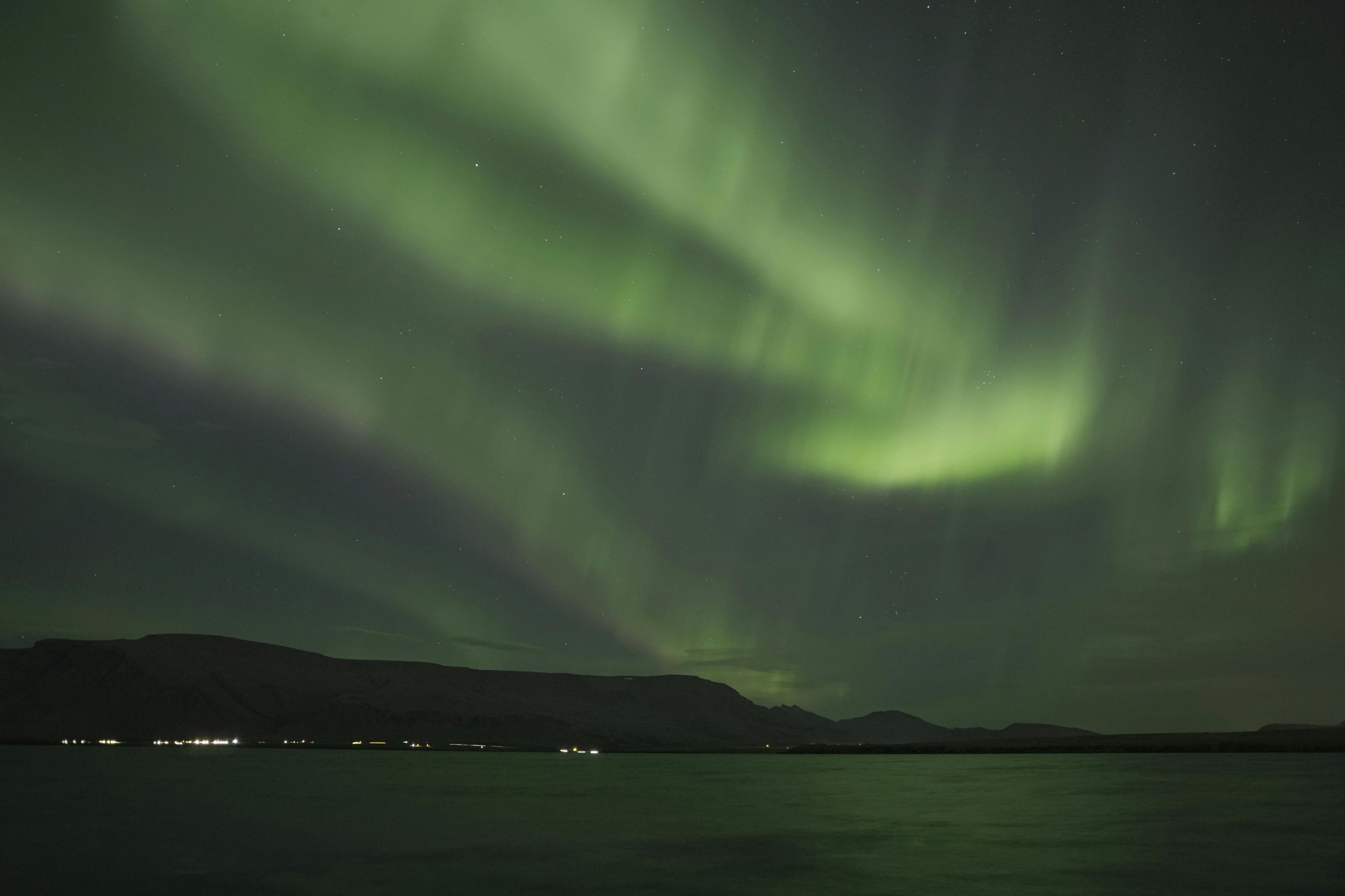 Green aurora borealis lights illuminate a dark, starry sky above distant hills and a calm body of water.