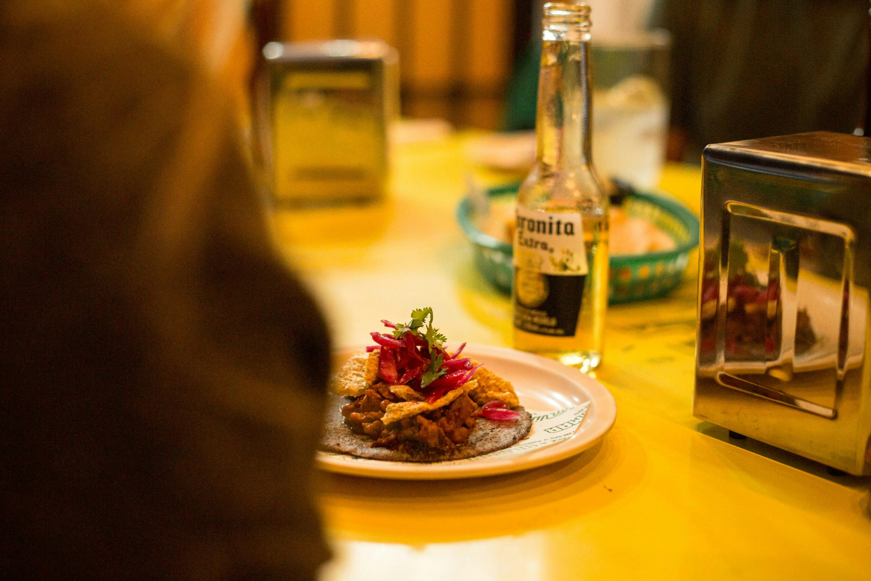 Un piatto di cibo con guarnizioni e una bottiglia di birra su un tavolo giallo, con altri oggetti e sfondo sfocato visibili.