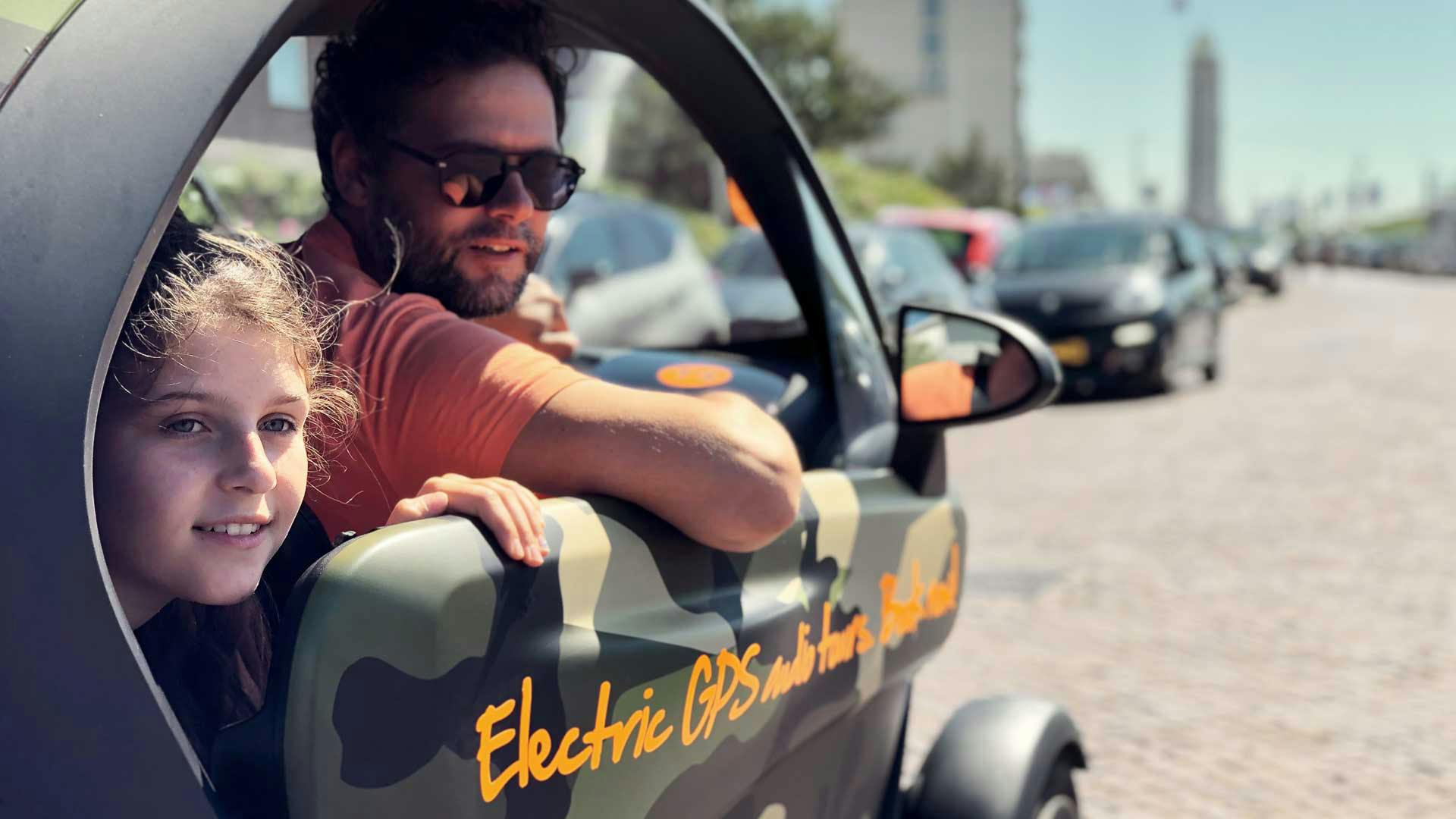 En mand iført solbriller og et barn kigger ud fra et vindue i et camo-mønstret køretøj med "Elektrisk GPS" skrevet på det.