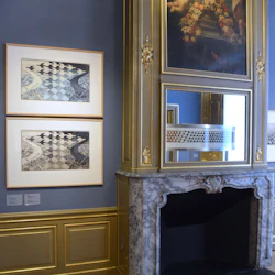 Ett rum med inramade konstverk, utsmyckad öppen spis och en stor spegel ovanför med gyllene accenter och ljust trägolv.