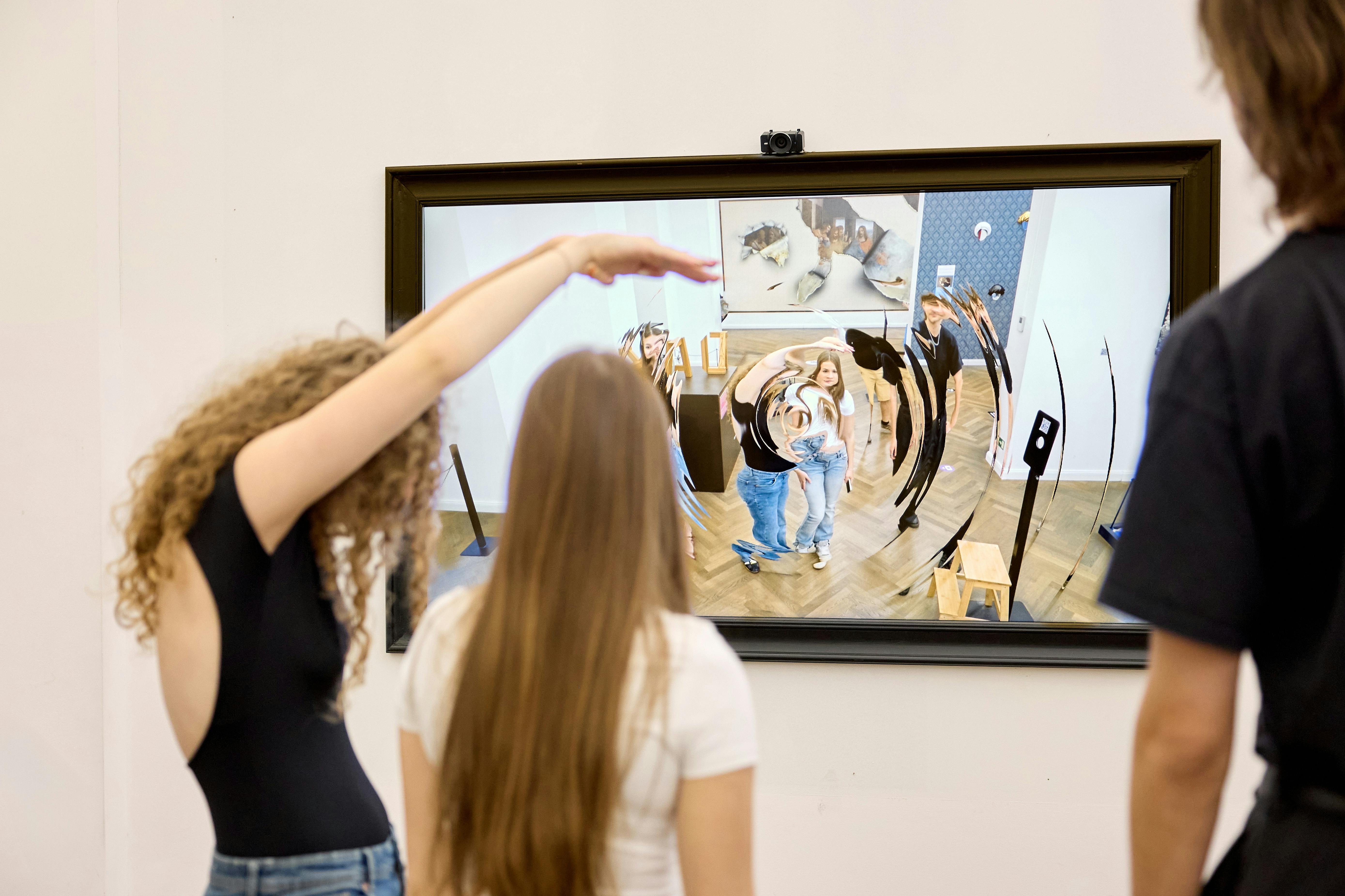 Folk står foran en skærm, der viser en forvrænget refleksion af dem i et kunstgalleri.