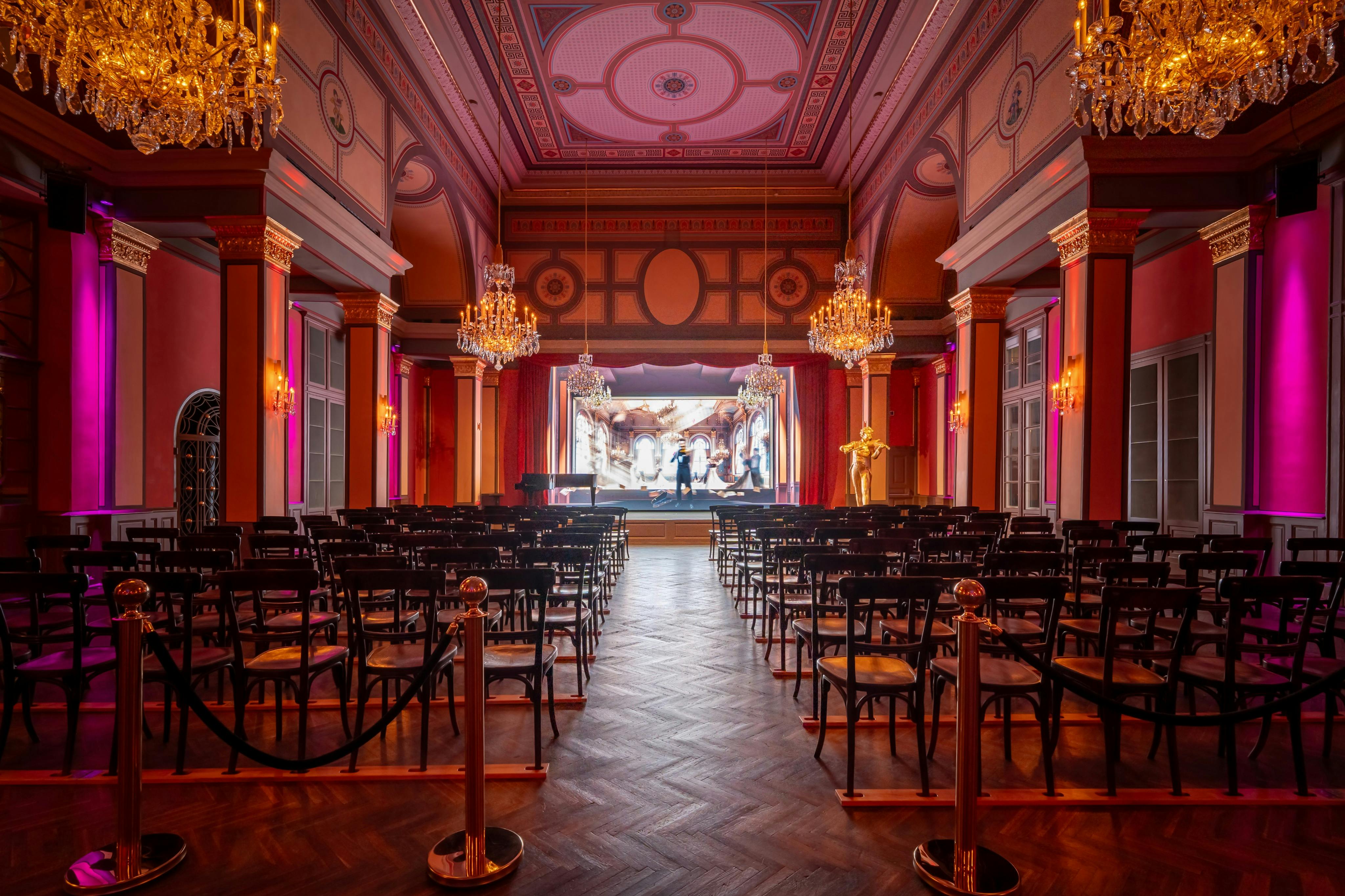 Ein großer Saal mit Stuhlreihen vor einer Bühne, von der Decke hängenden Kronleuchtern und kunstvollem Dekor.